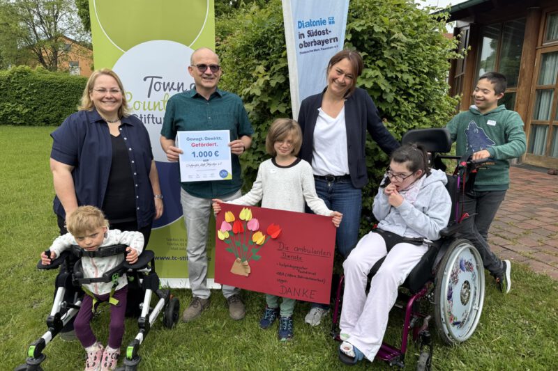 Große Freude bei der Spendenübergabe der Town & Country Stiftung an die Offene Behindertenarbeit Altötting: Kinder überreichen ein Plakat als Zeichen ihrer Freude über die Unterstützung für das Projekt „Andersgleich – Inklusion spielend erleben“. Stiftungsbotschafter Oliver Zahn übergibt die Spendenurkunde. (von links) Daniela Thalhammer (OBA Altötting), Oliver Zahn (Town & Country Stiftung) und Manuela Weiß (OBA Altötting).