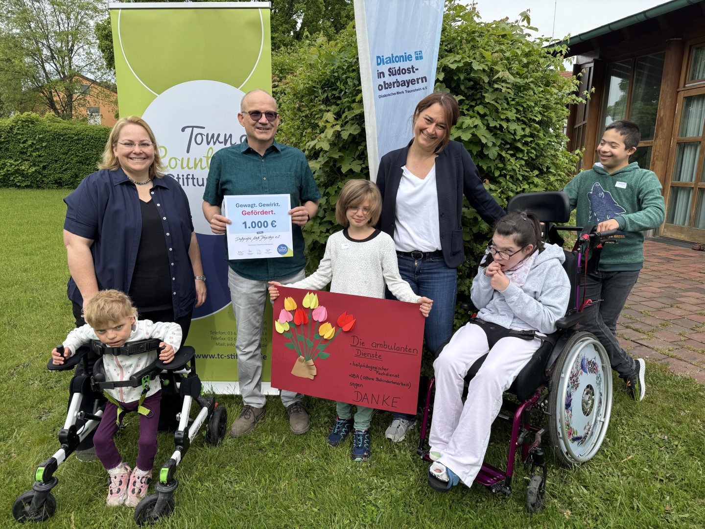 Große Freude bei der Spendenübergabe der Town & Country Stiftung an die Offene Behindertenarbeit Altötting: Kinder überreichen ein Plakat als Zeichen ihrer Freude über die Unterstützung für das Projekt „Andersgleich – Inklusion spielend erleben“. Stiftungsbotschafter Oliver Zahn übergibt die Spendenurkunde. (von links) Daniela Thalhammer (OBA Altötting), Oliver Zahn (Town & Country Stiftung) und Manuela Weiß (OBA Altötting).