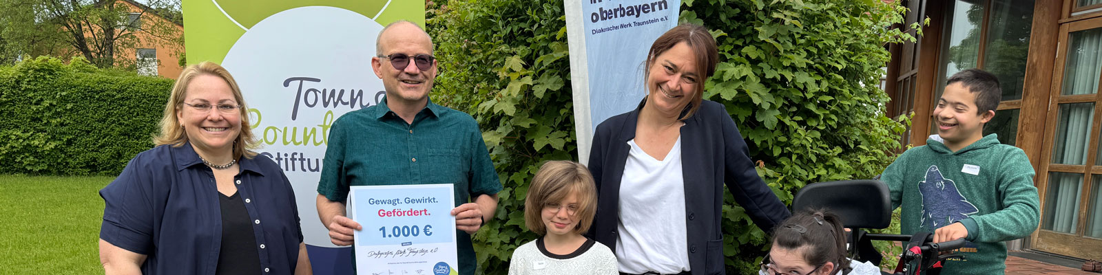 Große Freude bei der Spendenübergabe der Town & Country Stiftung an die Offene Behindertenarbeit Altötting: Kinder überreichen ein Plakat als Zeichen ihrer Freude über die Unterstützung für das Projekt „Andersgleich – Inklusion spielend erleben“. Stiftungsbotschafter Oliver Zahn übergibt die Spendenurkunde. (von links) Daniela Thalhammer (OBA Altötting), Oliver Zahn (Town & Country Stiftung) und Manuela Weiß (OBA Altötting).