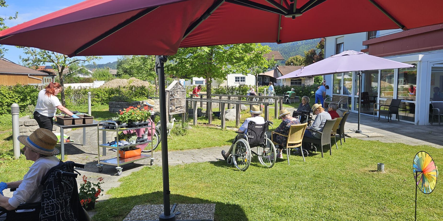 Bewohnerinnen und Bewohner bepflanzen gemeinsam mit Mitarbeitenden sommerliche Blumenkästen, andere Bewohner*innen sehen dabei zu und genießen das Sommerwetter.